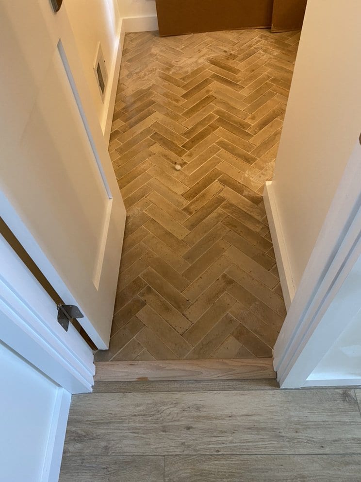 Finished hallway floor laid in a herringbone pattern of wood-effect planks, meeting a straight-laid floor at the threshold.